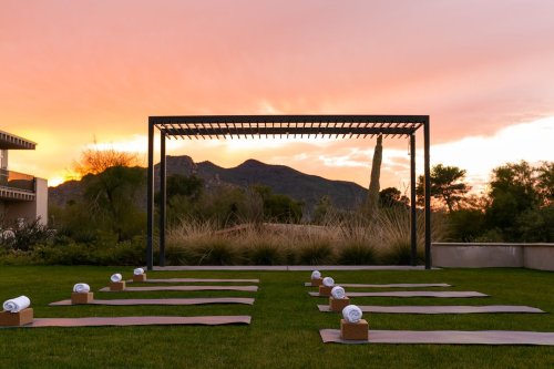 Yoga mats are arranged on grass with towels, framed by a sunset and mountains in the background.