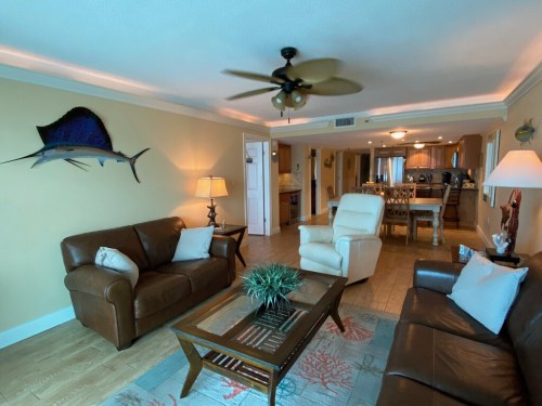 Cozy living room with brown leather sofas, a coffee table, ceiling fan, and a kitchen visible in the background.