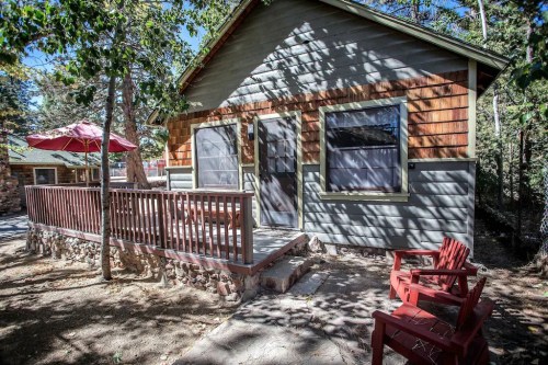 A cozy cabin with a wooden deck, surrounded by trees and a red umbrella, set in a natural outdoor setting.