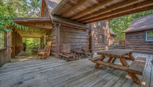 A rustic wooden cabin porch with rocking chairs and a picnic table, surrounded by trees.
