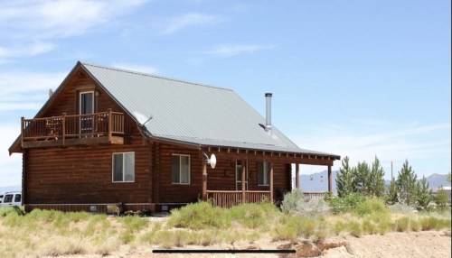 A rustic wooden cabin with a metal roof, surrounded by greenery and mountains under a clear blue sky.