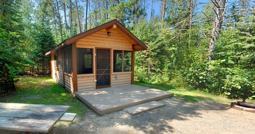 A cozy wooden cabin surrounded by trees, featuring a porch and a picnic table nearby.