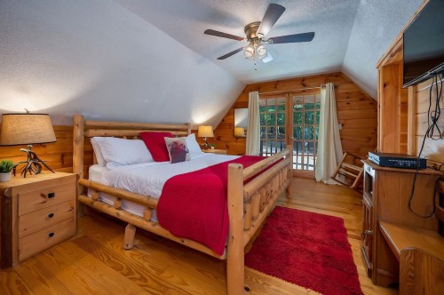 Cozy bedroom with wooden furniture, a red blanket, and a ceiling fan, featuring large windows and warm lighting.