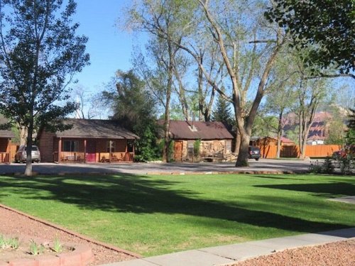 A grassy area with trees, featuring two rustic cabins and a gravel driveway under a clear blue sky.