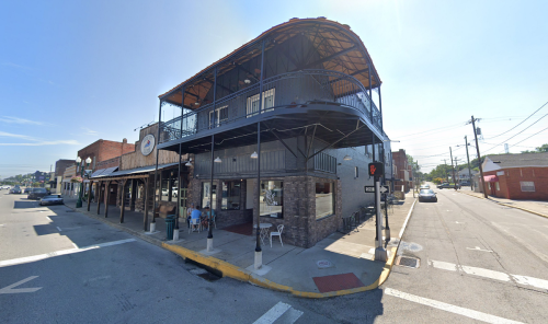 A two-story restaurant with a balcony, located at a street corner on a sunny day. Cars are parked nearby.