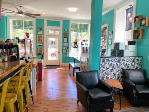 Brightly colored café interior with teal walls, wooden floors, bar seating, and cozy black chairs. Large windows let in natural light.