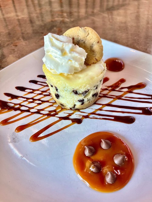 A dessert featuring a mini cheesecake topped with whipped cream, a cookie, and drizzled with caramel sauce.