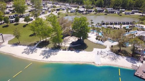Aerial view of a campground with RVs, a sandy beach, a pool area, and lush greenery surrounding a blue lake.