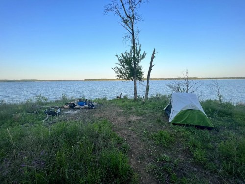 A tent set up by a calm lake, surrounded by grass and trees under a clear blue sky.