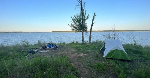 A serene lakeside campsite with a tent and gear set up on grassy land, surrounded by trees and calm water.