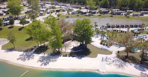 Aerial view of a campground with RVs, a pool area, and a sandy beach by a blue lake surrounded by trees.