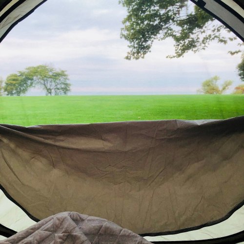 View from inside a tent, showcasing a green field and trees under a cloudy sky.