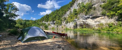 A tent by a calm river, surrounded by lush greenery and rocky cliffs under a blue sky with fluffy clouds.