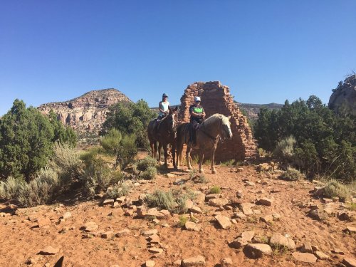 Two riders on horseback in a desert landscape with rocky formations and greenery under a clear blue sky.