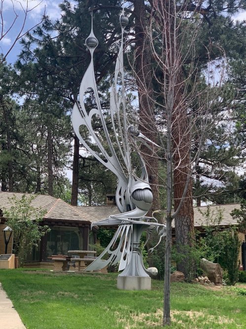 A large, abstract silver sculpture resembling a butterfly stands in a grassy area surrounded by trees.