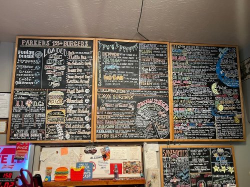 A chalkboard menu featuring various burgers, sandwiches, and drinks with colorful handwritten descriptions and prices.