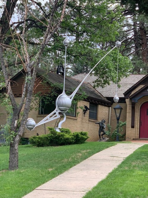 A unique, modern sculpture resembling a balance scale hangs from a tree in front of a house, surrounded by greenery.