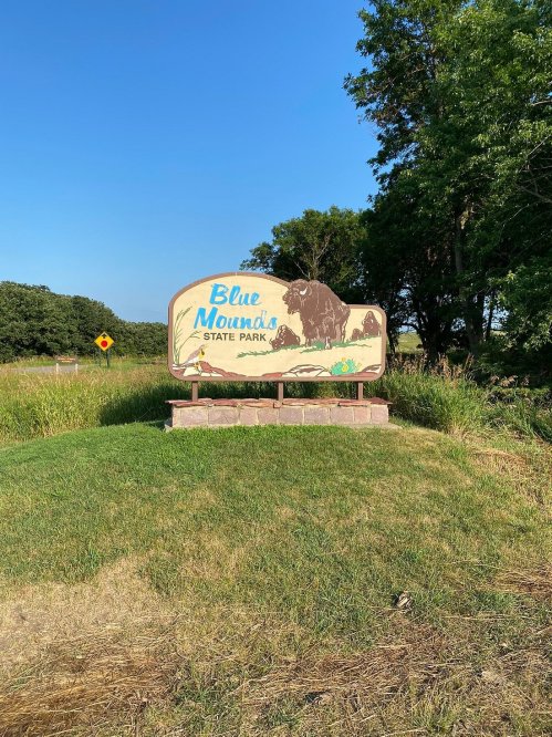 Sign for Blue Mountain State Park, featuring a bison graphic, surrounded by grass and trees under a clear blue sky.