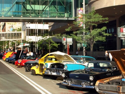 A row of classic cars parked along a city street, showcasing vibrant colors and open hoods under bright sunlight.