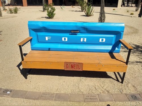 A bench made from a blue Ford truck tailgate, with wooden seating and a sign reading "O'Neill." Surrounded by gravel and plants.