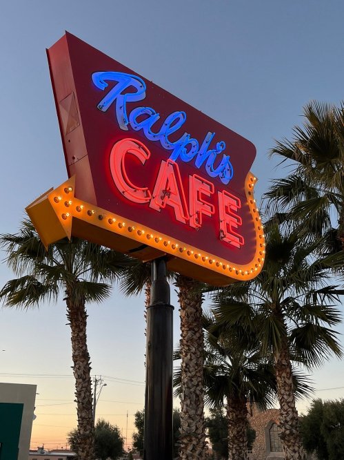 Neon sign for Ralph's Café illuminated at dusk, surrounded by palm trees.