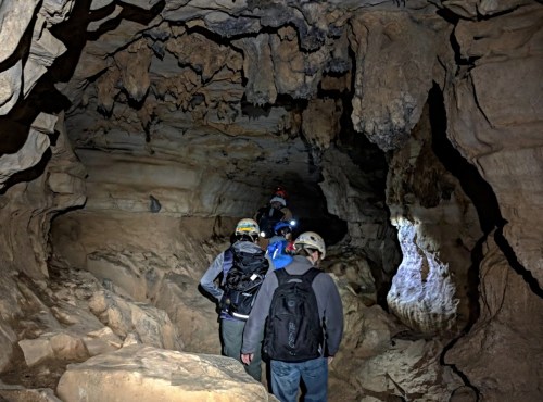 A group of people wearing helmets explores a dark cave with rocky walls and uneven terrain.