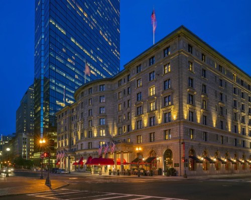 Historic hotel exterior illuminated at night, with modern skyscraper in the background and flags displayed.