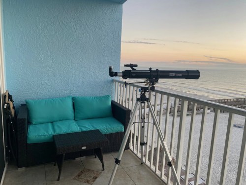 A telescope on a balcony overlooking the ocean at sunset, with a cozy seating area nearby.