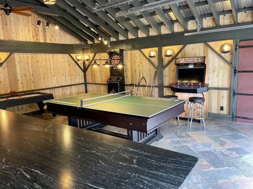 A rustic game room featuring a ping pong table, dartboard, and arcade machine, with wooden walls and a stone floor.