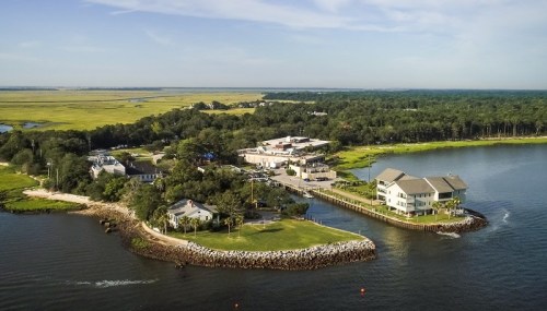 Aerial view of a coastal area featuring buildings, greenery, and water, with a serene landscape in the background.