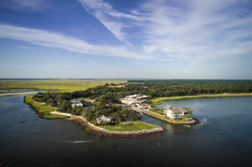 Aerial view of a coastal area with buildings, lush greenery, and calm waters under a blue sky with wispy clouds.