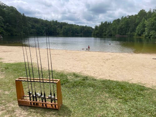 A sandy beach by a lake with fishing rods in a wooden holder and people swimming in the water in the background.