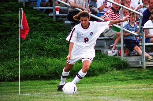 A female soccer player in a white uniform prepares to take a corner kick, with spectators in the background.