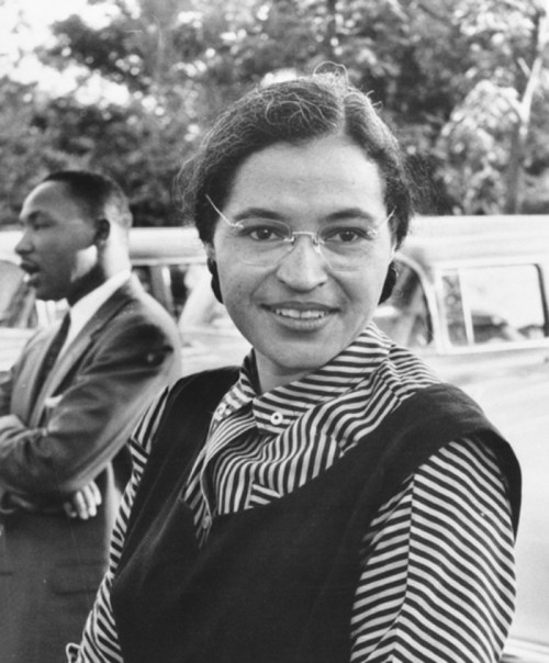 A black and white photo of a woman with glasses and a striped shirt, smiling, with a man in a suit in the background.