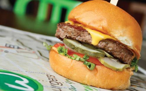 A close-up of a juicy cheeseburger with lettuce, tomato, pickles, and a toasted bun on a restaurant table.