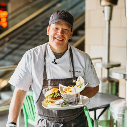 A chef in a cap smiles while holding a tray with a burger and fries in a modern restaurant setting.