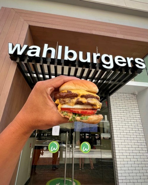 A hand holding a cheeseburger in front of a Wahlburgers restaurant sign.