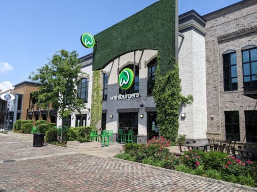 Exterior of a Wahlburgers restaurant with greenery, outdoor seating, and a brick pathway in a sunny setting.