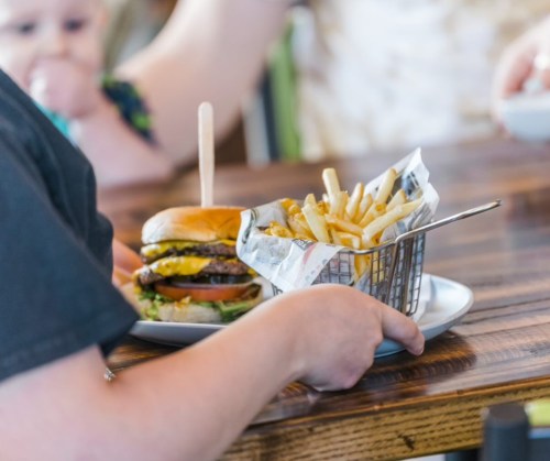 A person holds a plate with a burger and a basket of fries on a wooden table. A baby is partially visible in the background.