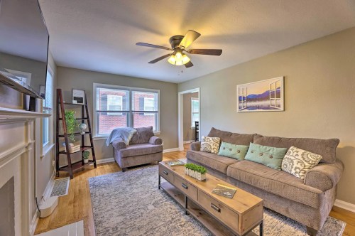 Cozy living room with a gray sofa, wooden coffee table, ceiling fan, and natural light from large windows.