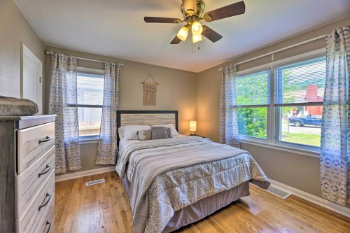 Cozy bedroom with a queen bed, ceiling fan, and large windows, featuring light curtains and wooden flooring.
