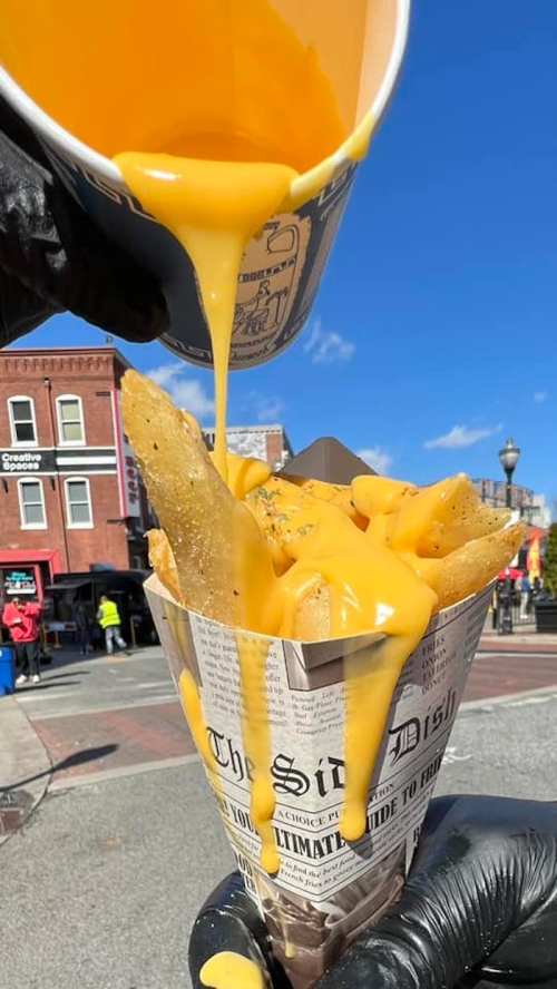 A hand pours melted cheese over a cone of crispy fries, set against a sunny outdoor backdrop.