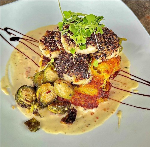 A plate featuring crispy scallops on cornbread, served with Brussels sprouts and a creamy sauce, garnished with microgreens.