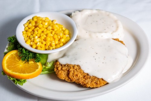 A plate with fried chicken smothered in gravy, mashed potatoes, corn, and a slice of orange on the side.