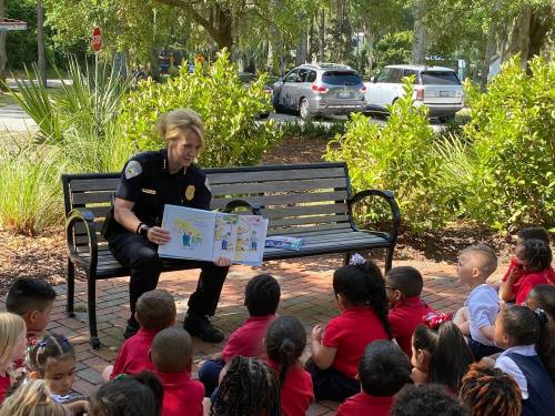 A police officer reads a story to a group of young children sitting on the ground in a park setting.