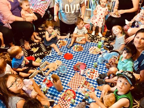 A group of children and adults gather on a blue checkered blanket for a picnic, surrounded by food and toys.