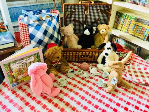 A tea party scene with stuffed animals around a table, a picnic basket, and a colorful book on a checkered tablecloth.