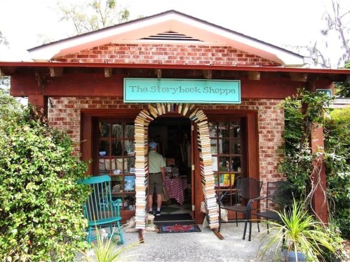 A quaint bookstore entrance with a brick facade, surrounded by greenery and featuring a colorful sign above the door.
