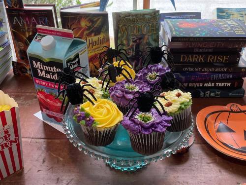 A glass cake stand holds colorful cupcakes topped with spiders, surrounded by books and a juice carton.