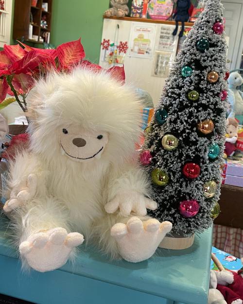 A fluffy white stuffed animal sits beside a small decorated Christmas tree with colorful ornaments.